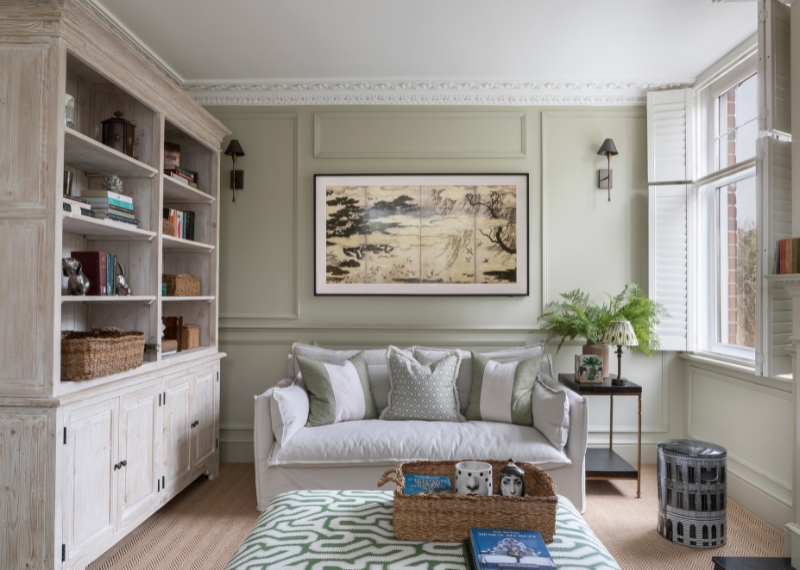 Soft sage green living room with classic wall panelling, bespoke shelving, and a neutral sofa layered with textured cushions and natural materials.
